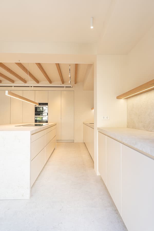 Architectural project - residential kitchen, white kitchen, stone surface, hidden storage, island countertop, LED strip, under-cabinet lighting, built-in appliances, culinary space, sustainable materials, wood, natural light, warm tones, eco design, beige, light wood, wood beams, timber beams, Scandinavian style, neutral palette, contemporary home, clean lines, white, Nordic design, inviting home, stone, plaster, open layout, open plan, integrated lighting, minimalist kitchen, modern kitchen, sleek design, functional layout, bright space, comfortable living, airy atmosphere, clutter-free, calming vibe, spacious feel, exposed ceiling