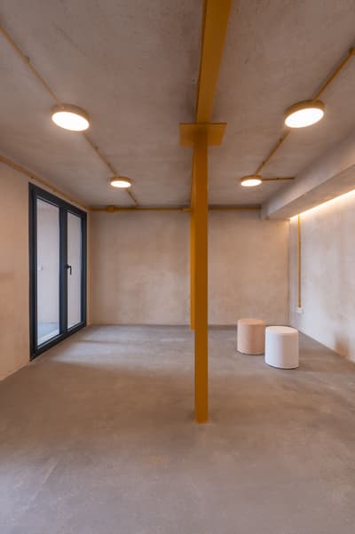 Architectural project - workshop space, exposed pipes, creative studio, unfinished look, modern renovation, urban chic, ceiling lights, simple seating, terrazzo floor, accent lighting, minimal design, raw materials, metal, natural light, yellow, exposed structure, grey, neutral tones, functional space, daylight, beige, concrete, industrial style, compact space, neutral palette, yellow accents, urban design, modern aesthetics, industrial chic, clean lines, plaster, open layout, multifunctional room, sleek design, plaster walls, concrete floor, contemporary vibe, industrial space, muted tones, minimalist interior