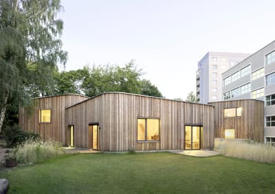 Architectural project - cozy interior, natural materials, wood, timber, glass, green, neutral tones, beige, light wood, low carbon design, daylight optimization, timber construction, honey wood, organic design, minimalist vibe, contemporary design, glass doors, sustainable building, wood panels, biophilic design, passive house, landscape integration, large windows, timber cladding, eco friendly home, inviting home, modern architecture, energy efficient, nature-inspired, garden view, wooden facade, residential building, simple lines, curved roof, suburban home, green space, minimal landscaping, urban context, warm lighting, sleek exterior, environmentally conscious, community housing, architectural harmony, urban garden, modern suburb