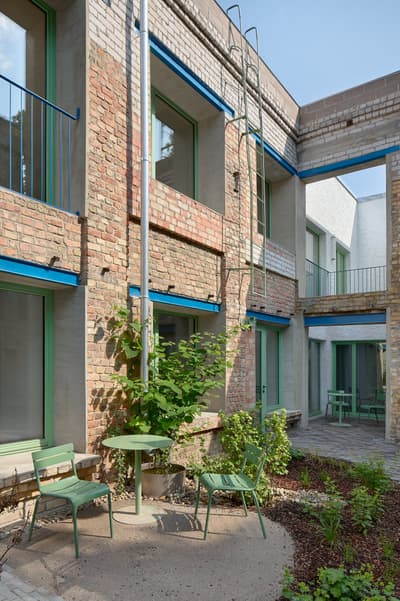 Architectural project - urban greenery, green accents, blue accents, creative reuse, metal railings, exposed brick, plant life, green elements, city courtyard, residential courtyard, open balcony, climbing plants, architectural renovation, recycled bricks, green chairs, minimalist, steel, outdoor space, natural light, warm tones, sustainable architecture, green, neutral tones, daylight, industrial vibe, brown, concrete, blue, urban oasis, contemporary design, exterior, adaptive reuse, earth tones, modern rustic, outdoor gathering, garden design, brick, eco friendly space, brick facade, textured walls, rustic aesthetic, garden seating, patio furniture, urban garden, courtyard, mixed materials, urban retreat, brick building, red