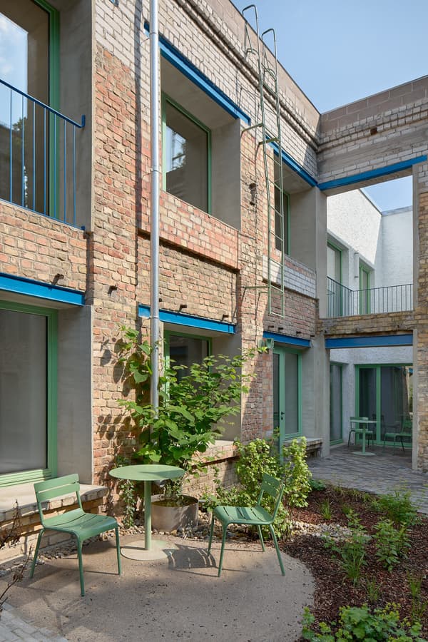 Architectural project - urban greenery, green accents, blue accents, creative reuse, metal railings, exposed brick, plant life, green elements, city courtyard, residential courtyard, open balcony, climbing plants, architectural renovation, recycled bricks, green chairs, minimalist, steel, outdoor space, natural light, warm tones, sustainable architecture, green, neutral tones, daylight, industrial vibe, brown, concrete, blue, urban oasis, contemporary design, exterior, adaptive reuse, earth tones, modern rustic, outdoor gathering, garden design, brick, eco friendly space, brick facade, textured walls, rustic aesthetic, garden seating, patio furniture, urban garden, courtyard, mixed materials, urban retreat, brick building, red