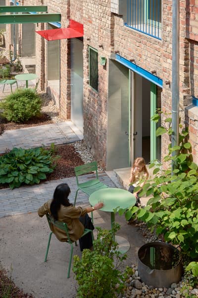 Architectural project - city garden, brick wall, lush plants, exposed brick, open door, vibrant accents, outdoor furniture, landscaped garden, urban courtyard, residential courtyard, small garden, green table, inner courtyard, metal canopy, colored canopy, green furniture, metal furniture, folding chair, modern courtyard, communal living, restful area, sustainable urban, warm brick, patio design, functional landscaping, natural materials, eco-friendly design, sustainable materials, metal, relaxed vibe, green, beige, brown, compact space, social space, gathering area, outdoor seating, indoor outdoor, inviting space, earthy tones, brick, casual outdoor, cozy corner, brick facade, greenery, outdoor design, garden seating, foliage, urban garden, family-friendly, urban retreat, urban living, pop of color, red, communal space