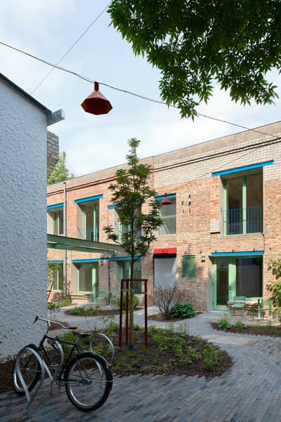 Architectural project - urban greenery, red accents, city garden, modern renovation, exposed brick, brick house, vibrant accents, social area, urban courtyard, open courtyard, garden courtyard, outdoor courtyard, green window frames, shared courtyard, bicycle parking, colorful frames, red lamp, outdoor lamp, light atmosphere, exterior brickwork, revitalized space, open gathering, natural materials, wood, steel, natural light, outdoor living, sustainable design, warm tones, eco design, community space, modern vibe, green, neutral tones, daylight, outdoor setting, concrete, multifunctional space, blue, urban oasis, rustic charm, industrial touch, public space, architectural details, inviting space, brick, cozy corner, brick facade, greenery, natural environment, garden seating, outdoor retreat, green landscaping, exterior space, red, communal space