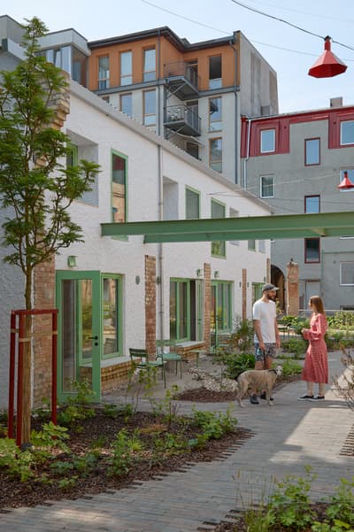 Architectural project - red accents, urban renewal, urban revitalization, vibrant community, mixed-use area, brickwork, roof terrace, urban texture, paved path, pastel green doors, white plaster, repurposed materials, eco district, green city, open-air living, shared garden, energy-efficient housing, neighborhood revitalization, city atmosphere, eco-friendly design, city living, cityscape, community space, sustainable architecture, green, urban oasis, contemporary design, social space, outdoor seating, color contrast, passive house, natural ventilation, modern facade, plaster, brick, greenery, modern living, light facade, urban garden, courtyard, daylight use, architectural contrast, connection to nature, residential block, mixed materials, urban housing, red