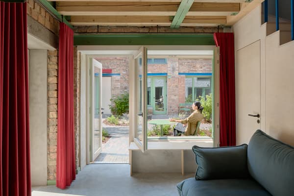 Architectural project - lounging area, brick wall, transitional space, indoor garden, exposed brick, polished concrete, indoor relaxation, soft furnishings, red curtains, vibrant curtains, patio access, plush sofa, cozy interior, wood, timber, relaxed vibe, natural light, warm tones, neutral tones, daylight, beige, concrete, blue, wood beams, timber beams, rustic modern, Scandinavian style, serene space, eco-friendly materials, modern rustic, eco home, sustainable home, indoor outdoor, Nordic style, outdoor view, comfortable home, casual living, open layout, open plan, inviting space, minimalist decor, brick, simple living, bright interior, concrete floor, airy feel, garden access, home living, red