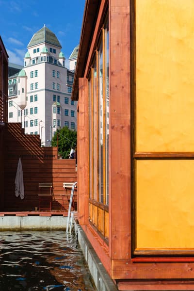 Architectural project - waterfront, vibrant facade, rooftop terrace, houseboat, water home, open windows, floating home, wooden siding, colorful architecture, balcony view, open water, city background, light reflection, minimal footprint, modern city life, water access, urban relaxation, city skyline, wood, timber, cityscape, outdoor space, yellow, glass, warm tones, eco design, contemporary style, beige, concrete, recreational space, modern design, urban oasis, honey wood, glass doors, vibrant colors, blue sky, green living, cozy outdoor, timber exterior, compact living, sky view, sustainable living, architectural contrast, urban retreat, urban living, red