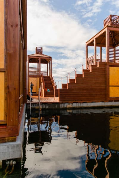Architectural project - floating structure, open deck, water reflection, floating platform, waterfront design, nautical style, outdoor stairs, raised platform, marina vibe, modern pier, water-based structure, harbor style, summer retreat, dockside feel, cloudscape, architectural interest, nautic theme, natural materials, wood, timber, passive design, warm tones, modular design, sustainable architecture, neutral tones, beige, brown, natural wood, open structure, natural setting, sunlight, timber construction, leisure space, contemporary design, seaside retreat, public space, gathering area, outdoor seating, lightweight structure, scenic view, wooden architecture, eco-friendly structure, sky view, environment integration, minimalism, timber steps