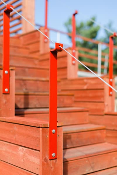 Architectural project - eco friendly material, outdoor access, red exterior, red stair, outdoor stair, red railing, wooden steps, open risers, exterior steps, exterior stair, modern railing, minimalist railing, vibrant outdoor, garden stair, patio steps, deck steps, wood grains, landscape feature, exterior architecture, garden connection, eye-catching design, deck area, natural materials, architectural detail, wood, timber, playful design, outdoor living, sustainable design, warm tones, neutral tones, beige, light wood, natural setting, rustic charm, contemporary design, warm wood, backyard feature, garden feature, casual outdoor, sunny day, timber steps, wooden stair, red