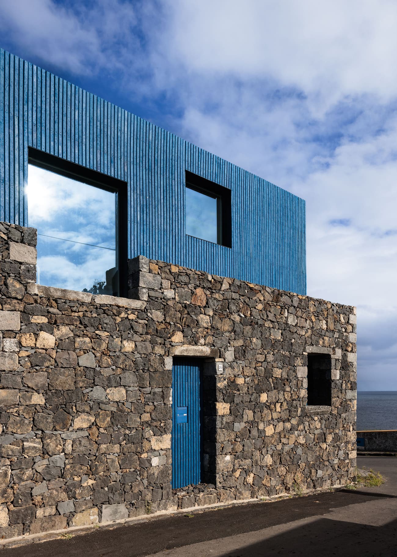 Architectural project - stone facade, natural stone, vertical lines, blue accents, blue cladding, ocean view, geometric windows, bold design, coastal house, dark stone, volcanic stone, basalt facade, ribbed cladding, seaside home, rugged texture, vibrant blue, horizontal lines, stacked layout, angular design, weathered look, natural elements, contrasting materials, sea view, cubic form, modern residence, oceanfront location, raw materials, outdoor space, sustainable design, minimalist design, eco-friendly materials, durable materials, large windows, modern architecture, earthy tones, contemporary house, textured facade, modern exterior, architectural rhythm, architectural contrast, exterior cladding, architectural geometry