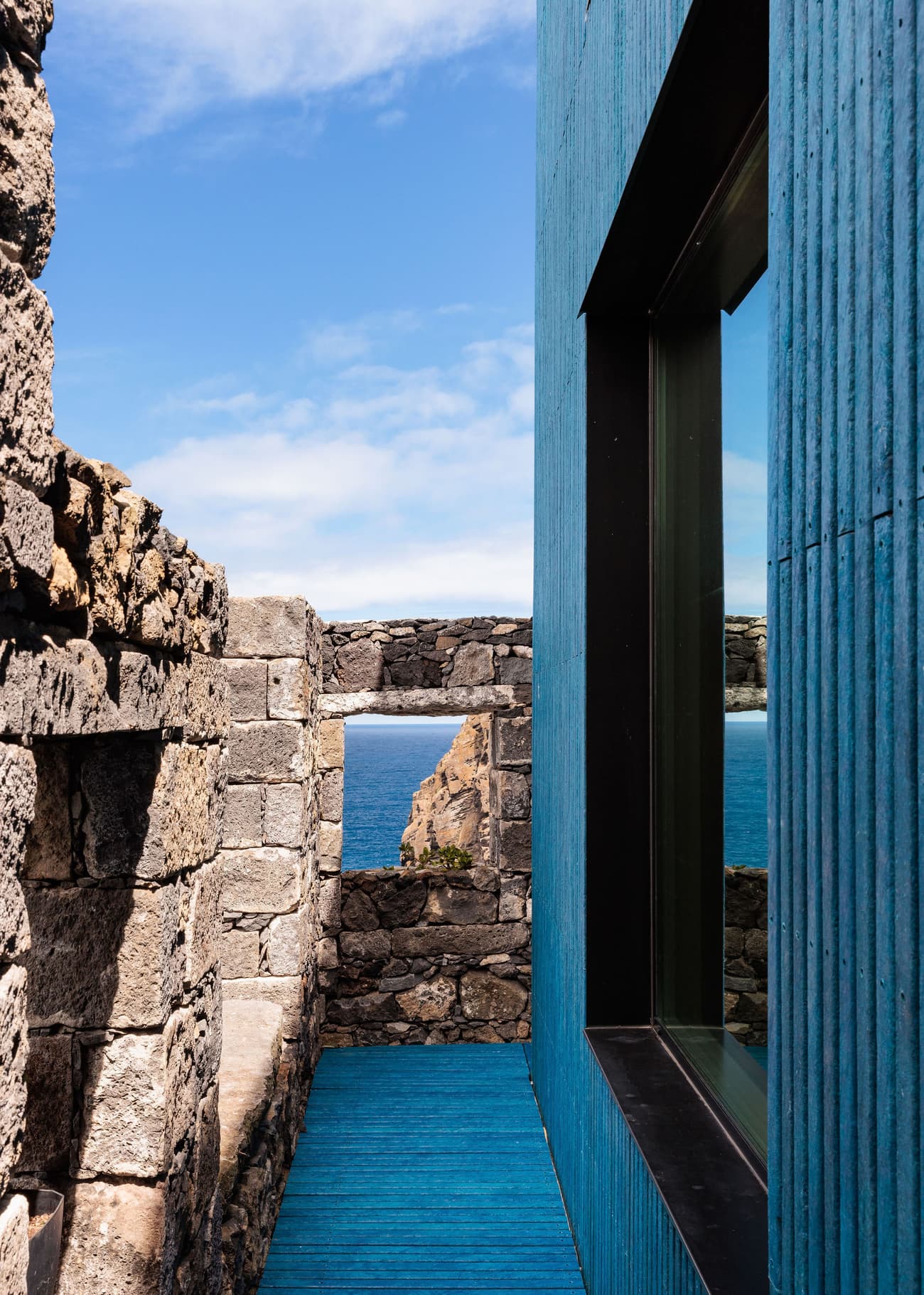 Architectural project - natural stone, blue cladding, blue facade, ocean view, vibrant facade, textured cladding, rustic stone, coastal view, island architecture, seaside architecture, bright blue, deep blue, external corridor, modern addition, contemporary extension, outdoor walkway, monochromatic scheme, cladding, vibrant exterior, sleek window, window opening, juxtaposition, ocean backdrop, architectural detail, outdoor space, natural light, contemporary architecture, contemporary style, grey, neutral tones, daylight, blue, modern design, rustic modern, minimalist style, modern rustic, stone, stone wall, minimalist exterior, textured facade, modern exterior, architectural contrast, stone texture