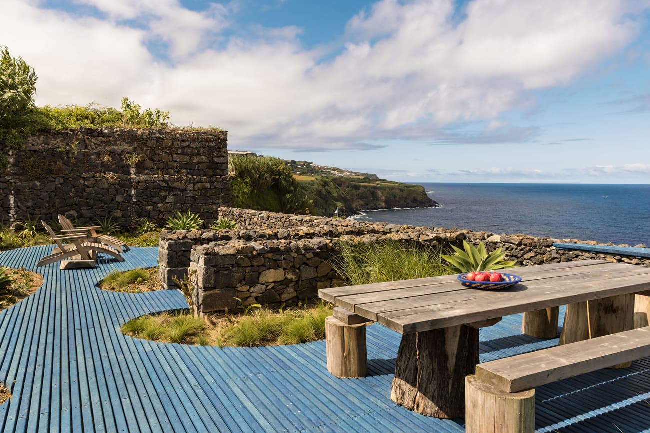 Architectural project - natural stone, outdoor lifestyle, retreat design, oceanfront, ocean view, coastal garden, organic landscape, open sky, coastal retreat, seaside view, outdoor lounge, stone walls, dry stone, picnic table, rustic bench, lush plants, wild garden, blue decking, nautical theme, getaway spot, sea breeze, stone architecture, rustic landscape, rugged finish, oceanfront living, rustic chic, wood, timber, beach house, coastal style, sustainable design, natural tones, grey, blue, eco friendly, natural textures, outdoor seating, earthy materials, stone, stonework, rustic style, wooden table, earthy tones, al fresco dining, rustic wall, relaxing space, peaceful retreat, timber deck, greenery, nature integration, vacation home, wooden elements, wooden deck, outdoor dining, nature blend, bohemian vibe