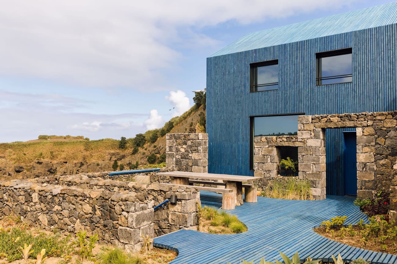 Architectural project - bold colors, blue facade, bright blue, stone walls, picnic table, wild garden, ribbed facade, garden patio, stone bench, lush scenery, coastal house, textured exterior, corrugated metal roof, ribbed roof, coastal vibe, cliffside house, minimalist garden, rustic, inviting outdoor, natural materials, metal, outdoor living, eco design, sustainable architecture, grey, neutral tones, exterior design, blue, natural setting, rustic modern, organic design, outdoor seating, natural landscape, modern rustic, scenic view, landscape integration, eco friendly home, stone, nature-inspired, grey stone, contemporary facade, garden path, modern style, open space, stone texture