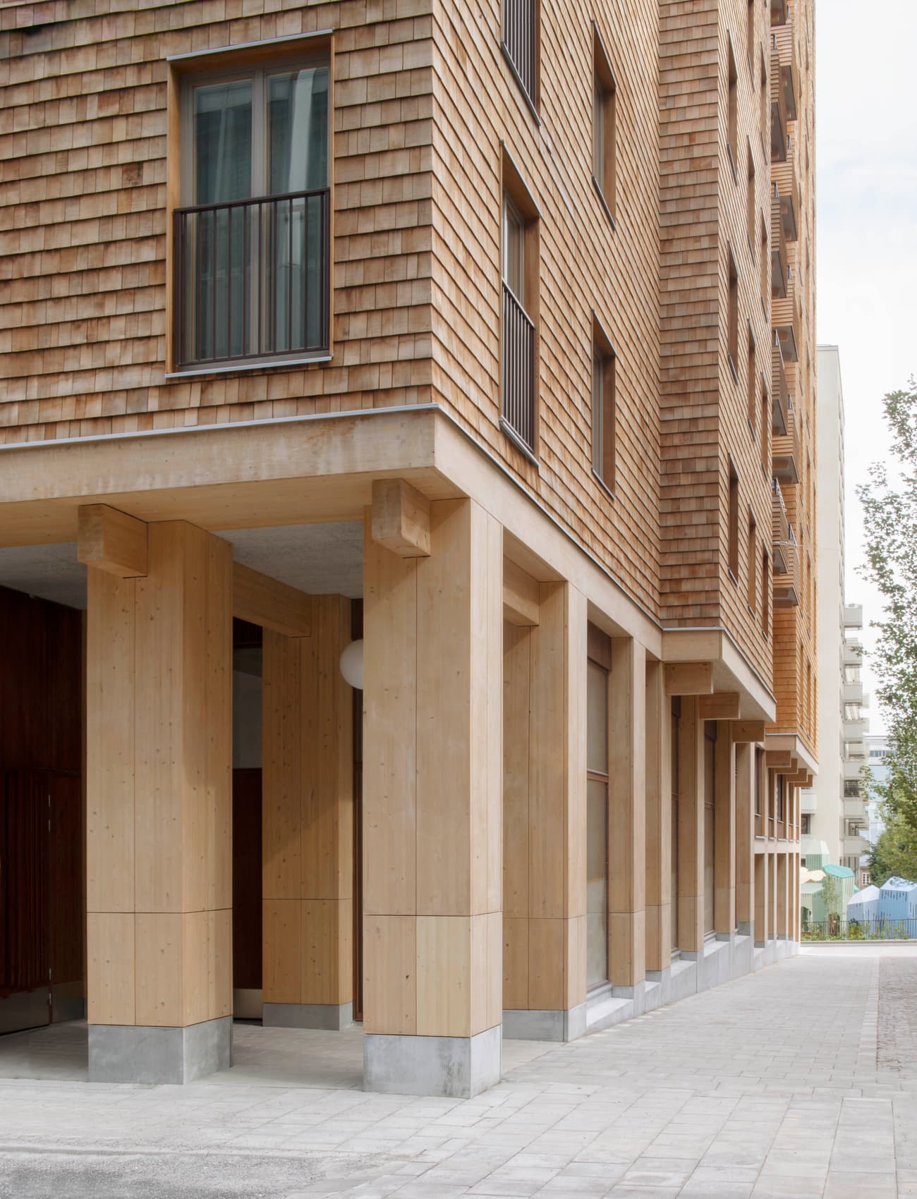 Architectural project - apartment block, apartment entrance, urban dwelling, urban residential, compact structure, textured exterior, communal living, natural materials, architectural detail, wood, timber, city living, passive design, sustainable design, warm tones, sustainable construction, urban landscape, beige, brown, concrete, light wood, daylight optimization, Scandinavian influence, minimalist design, timber detailing, eco building, wood panels, modern facade, timber cladding, modern architecture, Nordic style, cedar shingles, residential architecture, wooden facade, exterior wood, urban home, street view, contemporary building