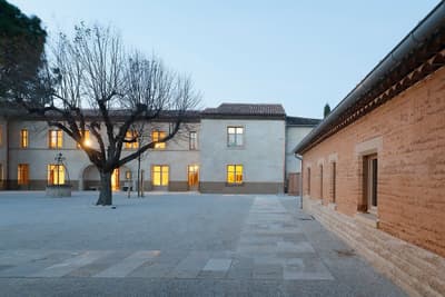 Architectural project - gathering place, stone walls, tiled roof, evening ambiance, outdoor lighting, symmetrical design, historic building, historic preservation, regional architecture, plaza, stone pathway, mature tree, heritage site, old building, cultural landmark, medieval style, village vibe, public square, conservation project, natural materials, outdoor space, passive design, warm tones, natural tones, sustainable construction, community space, open area, beige, low carbon design, eco friendly, urban oasis, serene atmosphere, calming space, neutral colors, tranquil setting, ecological design, natural landscape, earth tones, stone, earthy tones, pitched roof, traditional architecture, terracotta, rustic facade, textured walls, gravel path, sustainable facade, warm lighting, courtyard, light beige