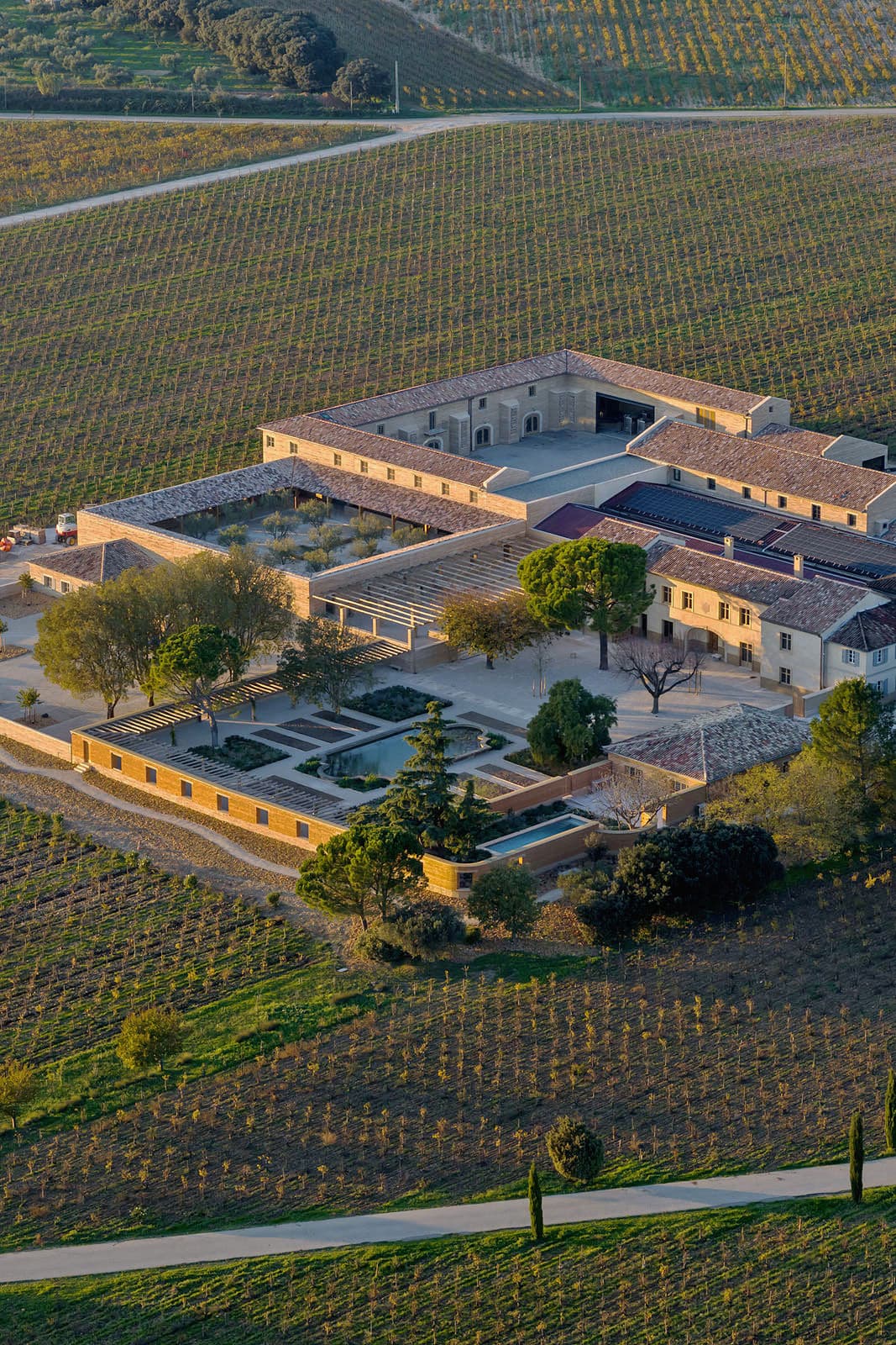 Architectural project - stone walls, tiled roof, historic building, terracotta roof, olive trees, classic design, vineyard estate, vineyard rows, sustainable agriculture, stone building, vineyard view, mediterranean architecture, rural estate, aerial shot, agricultural landscape, sprawling estate, farm estate, central courtyard, geometric layout, agricultural fields, eco-friendly farm, historic charm, estate grounds, green spaces, plantation, timeless elegance, heritage property, serene environment, green, beige, brown, rural architecture, rustic vibe, rustic charm, tranquil setting, earth tones, open landscape, scenic view, stone, traditional architecture, terracotta, countryside, landscape design, traditional style, countryside retreat, pastoral scene, courtyard