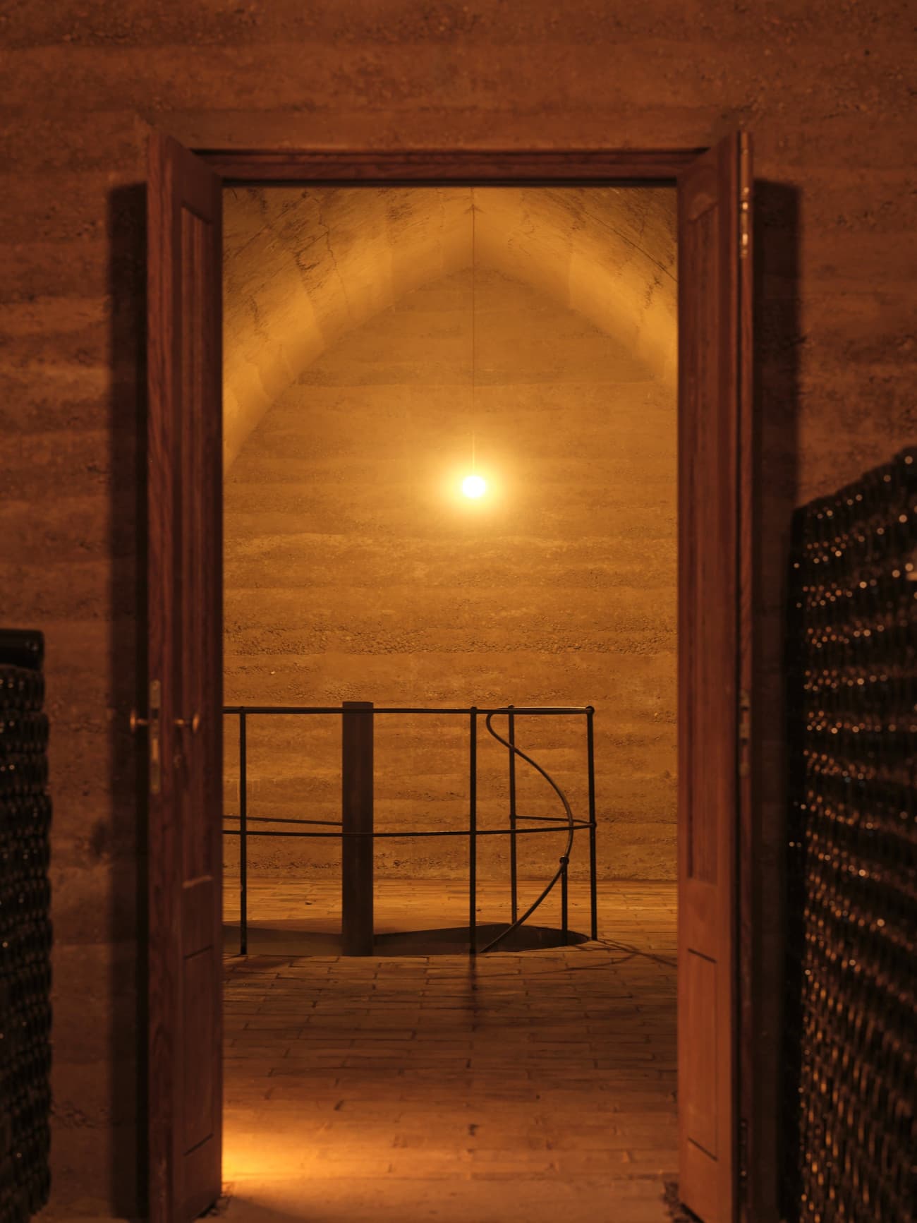 Architectural project - metal railing, soft glow, ambient light, rammed earth walls, muted palette, historic vibe, underground space, cozy lighting, earth tone walls, wine bottles, vintage elegance, secluded, private wine space, earthen finish, ceramic floor, heritage space, low light, timeless feel, brick floor, wine cellar, rustic cellar, arched ceiling, storage space, enclosed space, bottle storage, intimate space, yellow, warm tones, neutral tones, beige, brown, rammed earth, warm atmosphere, rustic design, natural texture, traditional style, black railing, rustic textures, warm lighting, earthy colors, wood door, artisanal touch, vaulted ceiling
