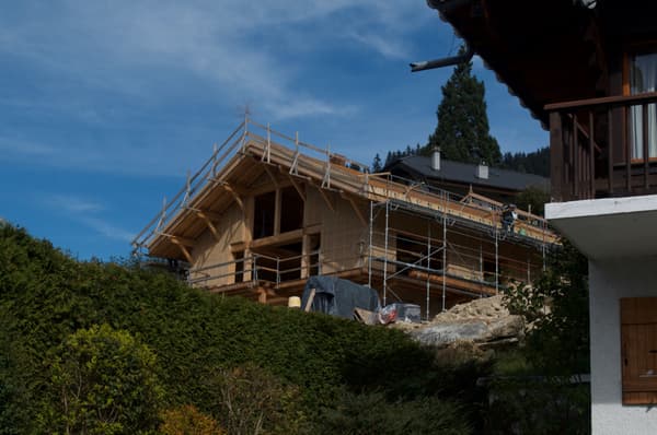 Architectural project - exterior structure, nature backdrop, vernacular style, scenic setting, new build, sustainable materials, wood, timber, scaffolding, metal, timber frame, green, construction site, work in progress, under construction, wood framework, beige, brown, timber facade, eco friendly, natural setting, wooden beams, timber construction, rural architecture, rustic charm, open framework, wood construction, construction details, building process, blue sky, green architecture, hillside house, landscape integration, harmonious design, countryside home, outdoor view, pitched roof, forest view, wooden cladding, building project, rural retreat
