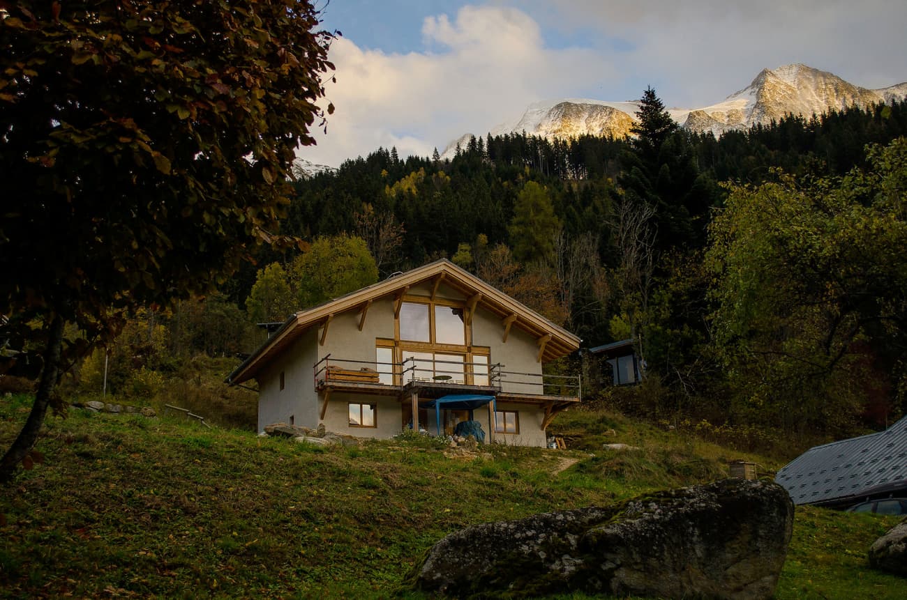 Architectural project - mountain retreat, forest backdrop, seasonal home, scenic location, rustic house, stucco, private getaway, wooden balcony, panoramic views, natural materials, wood, timber, natural light, outdoor living, passive design, warm tones, timber structure, beige, brown, wood beams, timber beams, cozy retreat, neutral palette, wood construction, natural landscape, earth tones, sustainable home, hillside house, large windows, inviting home, countryside home, stone, plaster, rural living, pitched roof, traditional design, tranquil atmosphere, gable roof, nature escape, serene setting, cottage style, cozy vibes, timber railing