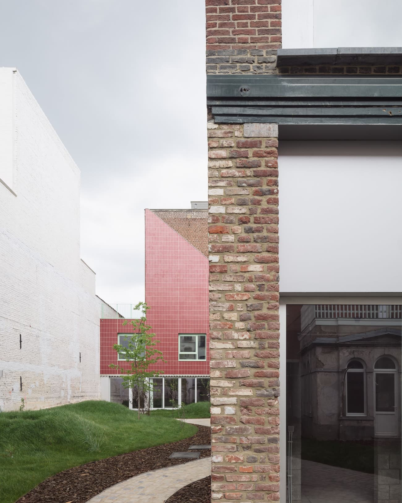 Architectural project - grass, light grey, red facade, brick wall, open sky, juxtaposition, brick exterior, landscaping, curved path, walking path, arched windows, historic touch, refurbished, contrast, external view, front yard, mixed material, textural contrast, minimalist, steel, cityscape, outdoor space, glass, contemporary architecture, urban landscape, neutral tones, concrete, industrial style, modern design, rustic charm, Scandinavian style, adaptive reuse, raw texture, white, brick, brick facade, heritage building, green lawn, renovation, smooth finish, brick building, street view, urban style, brick texture, urban vibe, red