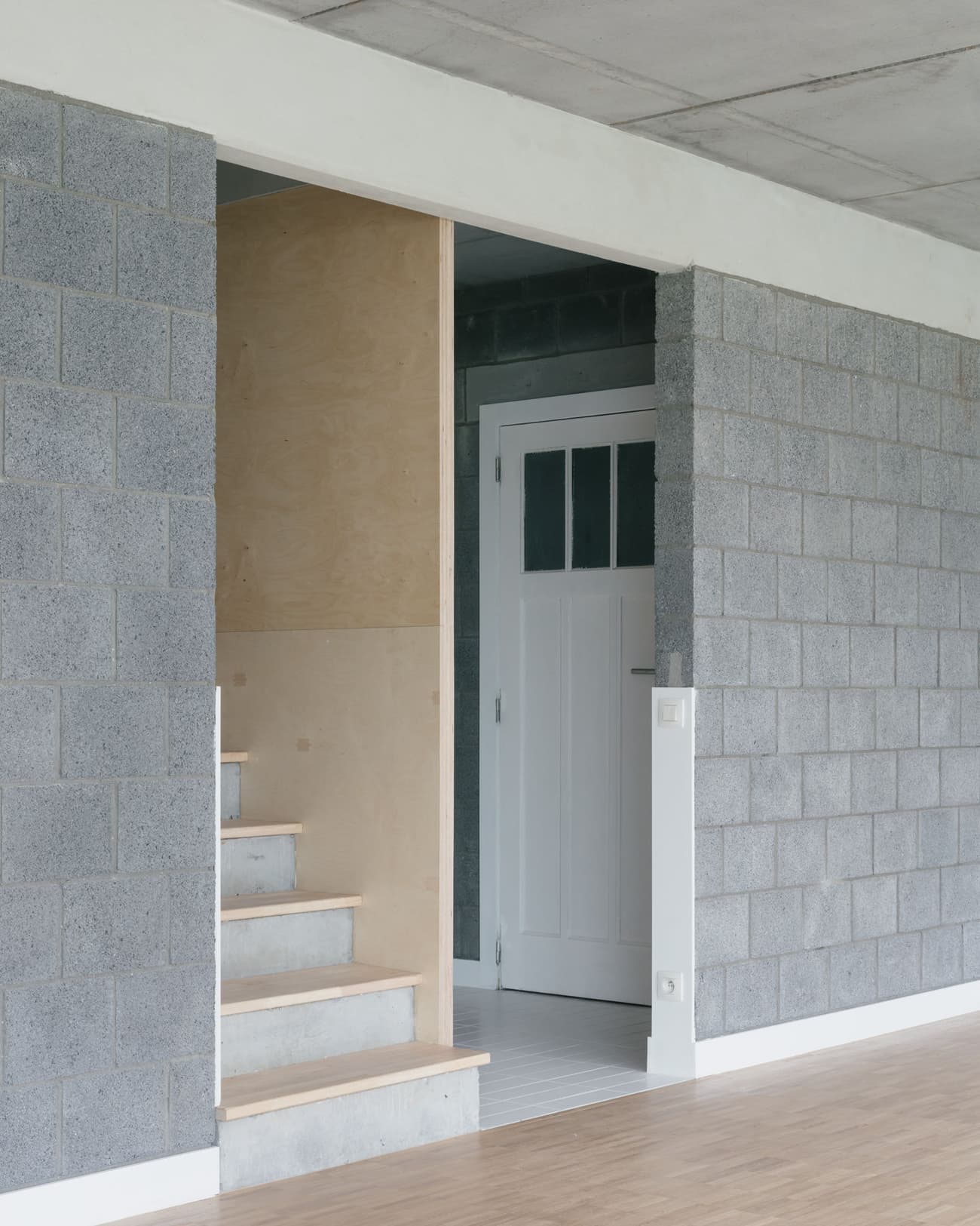Architectural project - contrast materials, concrete ceiling, transitional space, open door, compact layout, white trim, entrance, concrete block, cinder block, exposed block, modern minimal, plywood panel, grey wall, cozy entrance, home entry, warm minimalism, bauhaus influence, minimal design, architectural detail, wood, raw materials, passive design, sustainable design, eco design, grey, neutral tones, plywood, industrial vibe, concrete, light wood, natural wood, functional design, Scandinavian influence, architectural texture, industrial touch, minimalist style, neutral palette, contemporary interior, clean aesthetic, white, Nordic style, energy efficient, raw finish, simple lines, plywood stairs, timber steps, geometric form, stairwell, efficient space