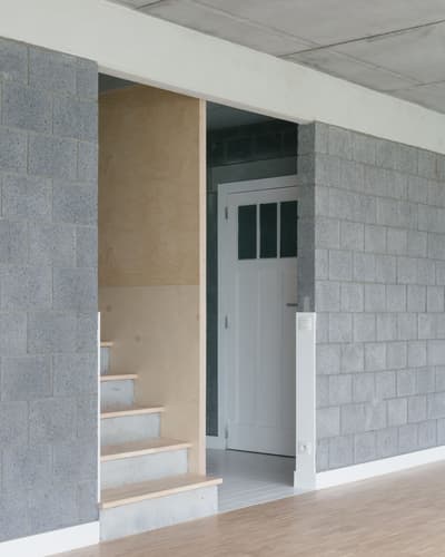 Architectural project - contrast materials, concrete ceiling, transitional space, open door, compact layout, white trim, entrance, concrete block, cinder block, exposed block, modern minimal, plywood panel, grey wall, cozy entrance, home entry, warm minimalism, bauhaus influence, minimal design, architectural detail, wood, raw materials, passive design, sustainable design, eco design, grey, neutral tones, plywood, industrial vibe, concrete, light wood, natural wood, functional design, Scandinavian influence, architectural texture, industrial touch, minimalist style, neutral palette, contemporary interior, clean aesthetic, white, Nordic style, energy efficient, raw finish, simple lines, plywood stairs, timber steps, geometric form, stairwell, efficient space