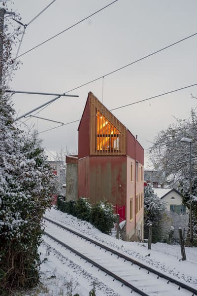 Architectural project - small house, warm glow, colorful exterior, red facade, city home, timber slats, suburban setting, urban house, asymmetrical design, natural materials, wood, timber, natural light, warm tones, eco design, contemporary style, evening light, beige, concrete, modern design, rustic charm, low carbon, sustainable home, passive house, modern architecture, energy efficient, plaster, sloped roof, pitched roof, textured walls, wooden facade, residential building, compact living, urban architecture, vertical cladding, architectural contrast, geometric form, urban retreat, red