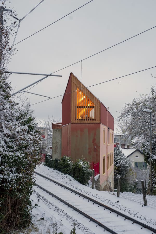 Architectural project - small house, warm glow, colorful exterior, red facade, city home, timber slats, suburban setting, urban house, asymmetrical design, natural materials, wood, timber, natural light, warm tones, eco design, contemporary style, evening light, beige, concrete, modern design, rustic charm, low carbon, sustainable home, passive house, modern architecture, energy efficient, plaster, sloped roof, pitched roof, textured walls, wooden facade, residential building, compact living, urban architecture, vertical cladding, architectural contrast, geometric form, urban retreat, red