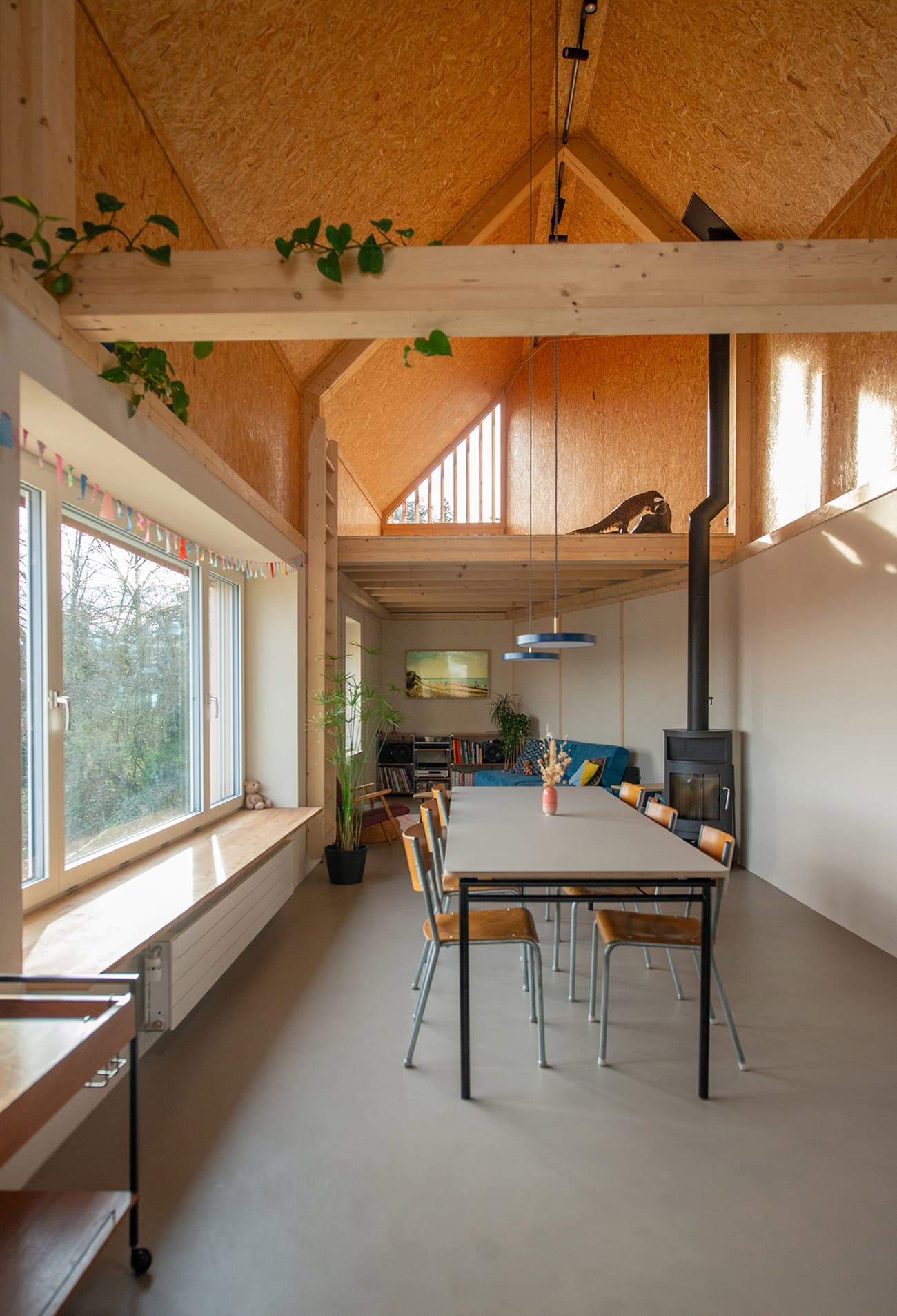 Architectural project - airy interior, modern dining, wooden chairs, dining chairs, cozy dining, potted plants, stylish home, minimalist table, natural materials, wood, timber, relaxed vibe, natural light, rustic touch, sustainable design, warm tones, grey, neutral tones, exposed beams, daylight, plywood, beige, light wood, golden, eco friendly, wooden beams, simple design, contemporary interior, passive house, large windows, Nordic style, energy efficient, timber ceiling, open layout, open plan, dining table, inviting space, family gathering, sloped roof, pitched roof, nature connection, greenery, Scandinavian design, modern living, wood ceiling, organic textures, pendant lamp, indoor plants, bright room, comfortable space
