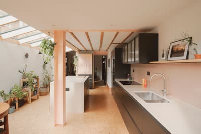 Architectural project - breakfast bar, indoor garden, integrated appliances, cork, open kitchen, black cabinets, white sink, sustainable materials, pink, wood, timber, natural light, glass, warm tones, neutral tones, daylight, beige, sunlight, timber beams, contemporary design, minimalist style, calming space, black, neutral colors, vibrant space, biophilic design, eco friendly home, inviting home, cozy kitchen, family kitchen, home kitchen, warm atmosphere, residential design, bright interior, modern kitchen, sleek design, kitchen island, wood ceiling, sustainable living, open shelving, skylight, airy kitchen, indoor plants