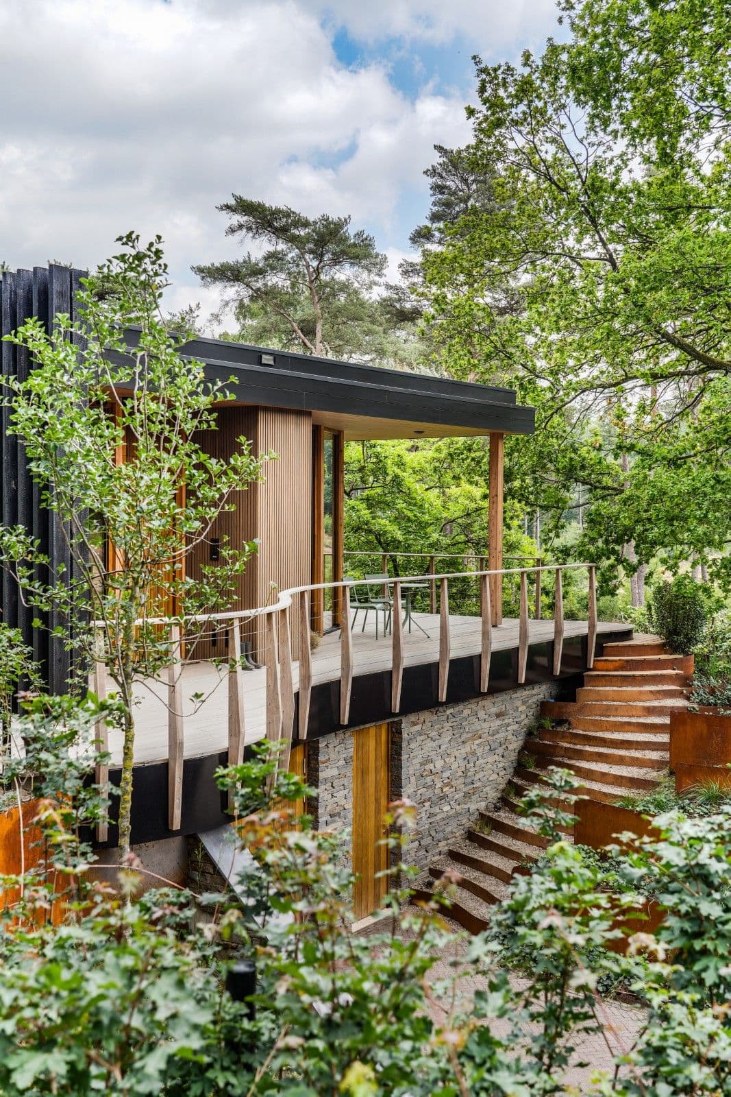 Architectural project - natural retreat, rustic elegance, zen retreat, woodland escape, tranquil design, elevated terrace, hillside home, treehouse design, layered landscaping, eco lodge, architectural staircase, weathered steel, corten stair, open shelter, roofed terrace, shaded deck, blending nature, lush environment, natural materials, timber, steel, outdoor living, glass, warm tones, green, neutral tones, brown, timber facade, minimalist design, black, biophilic design, landscape integration, eco friendly home, earthy materials, stone base, stone, rustic cabin, woodland setting, stone wall, nature retreat, forest view, peaceful retreat, timber deck, greenery, garden view, sustainable cabin, immersive nature, outdoor deck, wooden deck, residential, timber railing, vertical slats, modern cabin