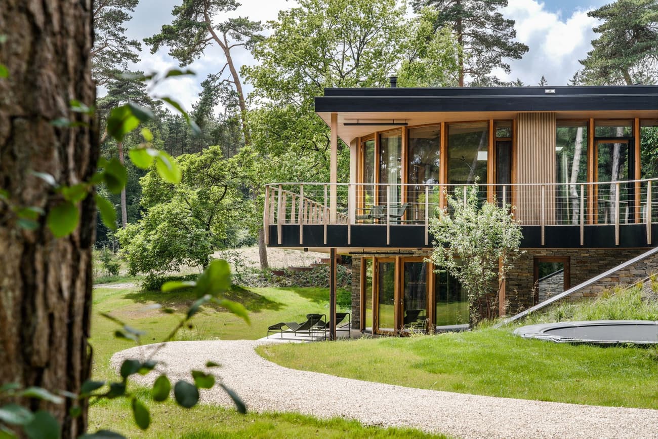 Architectural project - scenic location, earthy design, lush landscape, wooden balcony, curved facade, sloping site, family retreat, tranquil home, circular driveway, trampoline, serene vibe, natural materials, wood, timber, raw materials, outdoor space, outdoor living, glass, warm tones, eco design, neutral tones, daylight, recreational area, beige, light wood, low carbon design, eco friendly, natural setting, honey wood, peaceful setting, contemporary design, Scandinavian style, cozy retreat, minimalist design, sustainable home, passive house, clean lines, large windows, indoor outdoor, timber cladding, modern architecture, wood house, timber house, stone, open layout, inviting space, green home, nature retreat, garden view, gravel path, forest home, bright interior, nordic influence, glass facade, modern retreat, wood finish