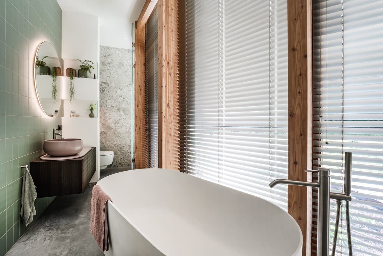 Architectural project - silver, polished concrete floor, green tiles, contemporary bathroom, round mirror, circular mirror, spa-like bathroom, freestanding tub, oval bathtub, white bathtub, green walls, wall-mounted sink, floating vanity, bathroom plants, wood blinds, privacy blinds, minimalist faucet, zen bathroom, modern finish, luxurious bathroom, architectural bathroom, eco-friendly design, wood, timber, metal, natural light, sustainable design, grey, green, daylight, concrete, calm ambiance, soothing tones, timber detailing, biophilic design, white, textured wall, airy space, earthy tones, organic materials, tranquil vibe, relaxing space, grey floor, bright interior, sleek faucet, soft colors, wood paneling, modern bathroom, ceramic, indoor plants