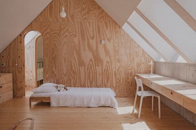 Architectural project - minimalist home, plywood walls, airy room, attic bedroom, white bedding, simple bed, built-in desk, minimalist desk, space optimization, warm interior, eco-friendly design, sustainable materials, timber, relaxed vibe, natural light, daylight, plywood, light wood, natural wood, modern aesthetic, Scandinavian influence, honey wood, modern simplicity, cozy retreat, neutral palette, serene space, timber walls, wood floor, timber elements, Nordic design, beige tones, small space, compact design, inviting space, wood texture, wooden flooring, peaceful vibe, bright interior, calm atmosphere, cozy home, sustainable living, minimalist bedroom, sloped ceiling, skylight, vaulted ceiling
