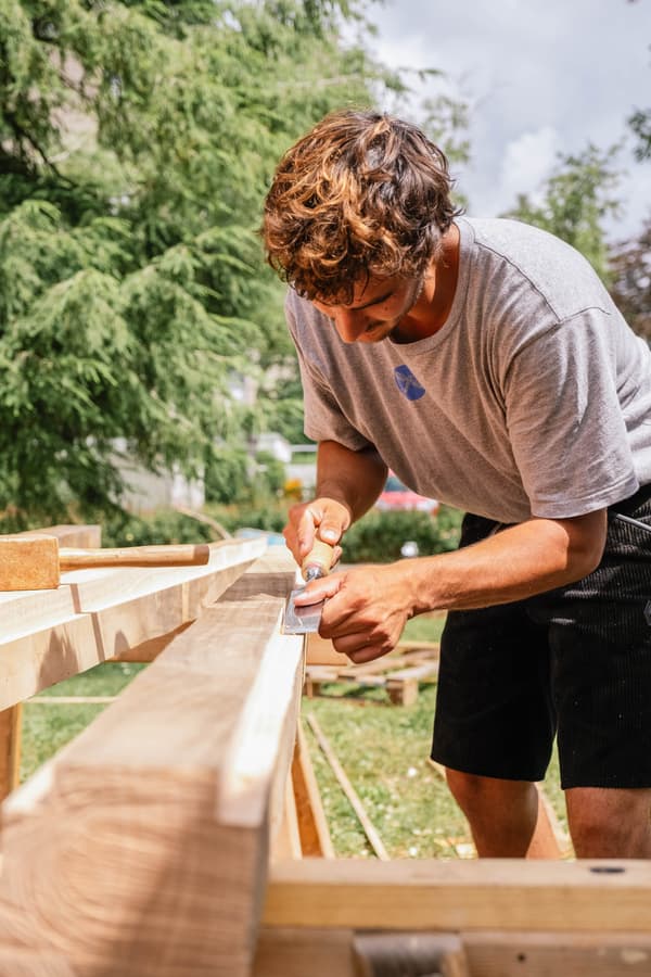 Architectural project - DIY project, handmade, outdoor workshop, skilled labor, manual labor, timber beam, woodworking, traditional technique, rustic construction, artisan, natural materials, wood, timber, outdoor space, timber frame, green building, construction site, carpentry, beige, brown, light wood, natural setting, eco-friendly construction, building process, wood structure, sustainable building, woodwork, earthy tones, natural texture, craftsmanship, construction