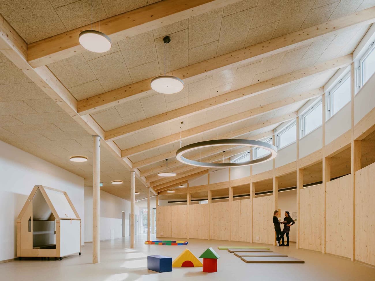 Architectural project - open ceiling, muted colors, playful area, cream, acoustic panels, soft lighting, wood, timber, warm tones, eco design, neutral tones, timber structure, exposed beams, beige, Scandinavian style, neutral palette, minimalist design, gathering area, community center, creative space, light tones, sustainable building, Nordic design, open plan, inviting space, wooden architecture, child friendly, wooden flooring, bright interior, educational space, modern interior, play area, family-friendly, versatile space