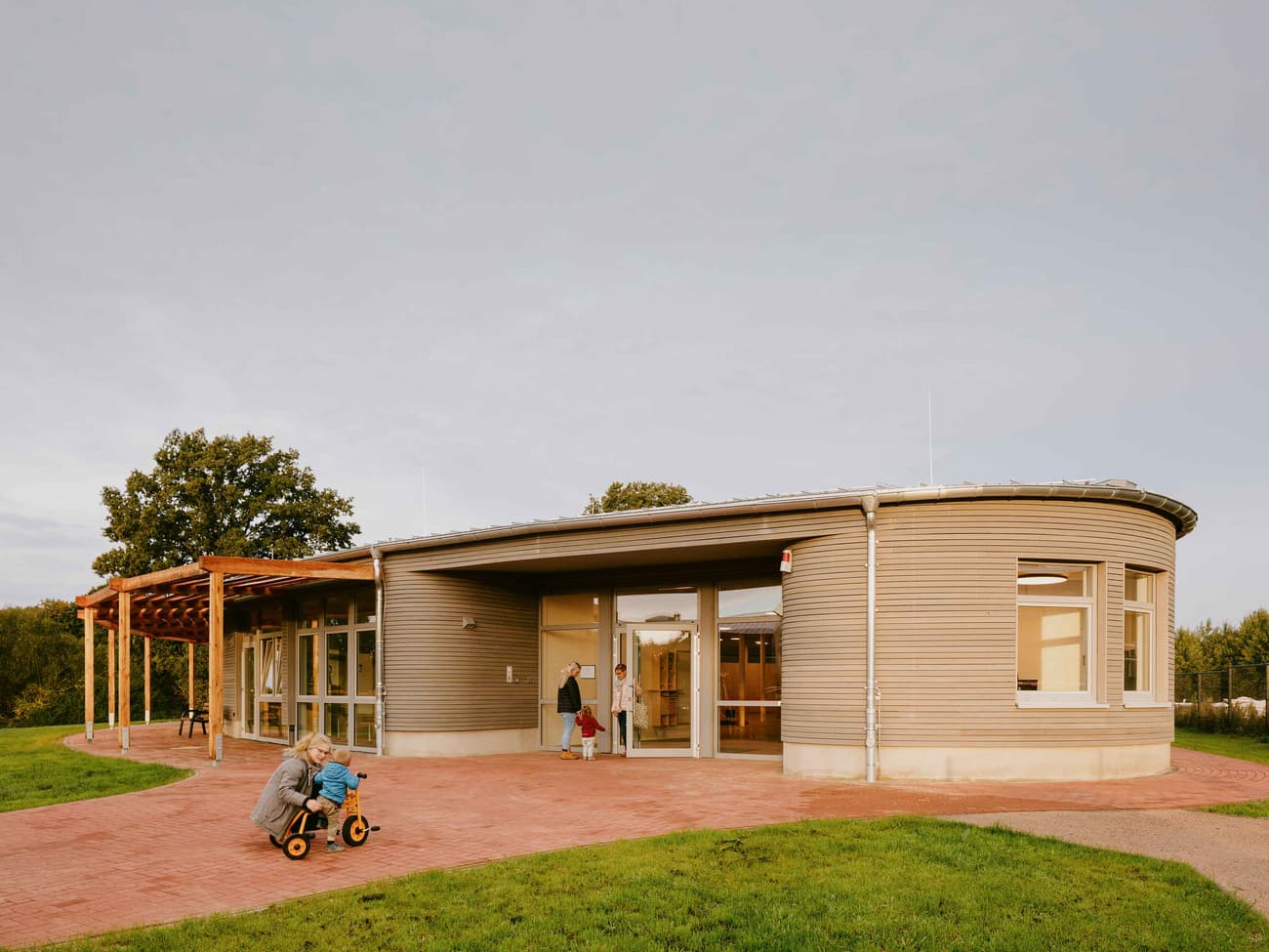 Architectural project - gray, metal siding, public gathering, welcoming entrance, wooden pergola, modern community, brick pathway, inclusive design, open entrance, natural materials, eco-friendly design, wood, metal, outdoor space, natural light, glass, eco design, public use, contemporary style, sustainable architecture, green, beige, timber facade, low carbon design, daylight optimization, modern design, community hub, community center, social hub, sustainable building, circular design, earth tones, passive house, corrugated metal, landscape integration, wood cladding, harmonious design, energy efficient, inviting space, outdoor gathering, child friendly, eco architecture, green surroundings, curved wall, public building, outdoor play, family-friendly