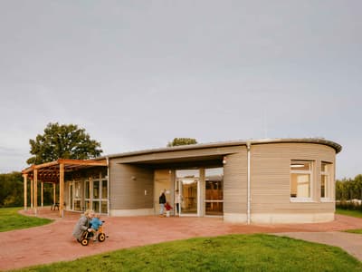 Architectural project - gray, metal siding, public gathering, welcoming entrance, wooden pergola, modern community, brick pathway, inclusive design, open entrance, natural materials, eco-friendly design, wood, metal, outdoor space, natural light, glass, eco design, public use, contemporary style, sustainable architecture, green, beige, timber facade, low carbon design, daylight optimization, modern design, community hub, community center, social hub, sustainable building, circular design, earth tones, passive house, corrugated metal, landscape integration, wood cladding, harmonious design, energy efficient, inviting space, outdoor gathering, child friendly, eco architecture, green surroundings, curved wall, public building, outdoor play, family-friendly