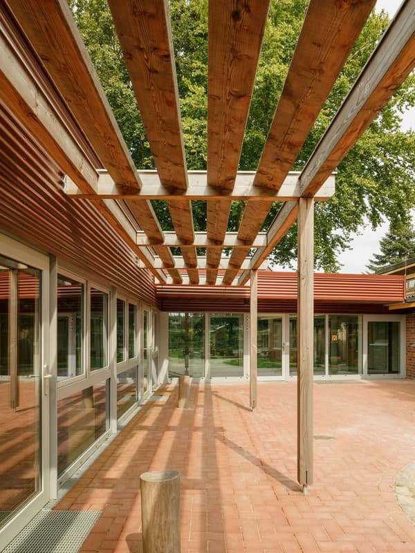 Architectural project - linear design, pergola, brick flooring, communal area, tree shade, natural materials, eco-friendly design, wood, timber, outdoor space, natural light, outdoor living, passive design, glass, warm tones, sustainable construction, community space, contemporary style, neutral tones, daylight, beige, brown, wooden beams, simple design, timber beams, minimalist design, social space, gathering area, glass doors, adaptive reuse, biophilic design, clean lines, modern architecture, inviting space, warm atmosphere, terracotta, brick, greenery, exterior view, tree integration, open air, sliding doors, architectural harmony, courtyard, terracotta floor, red