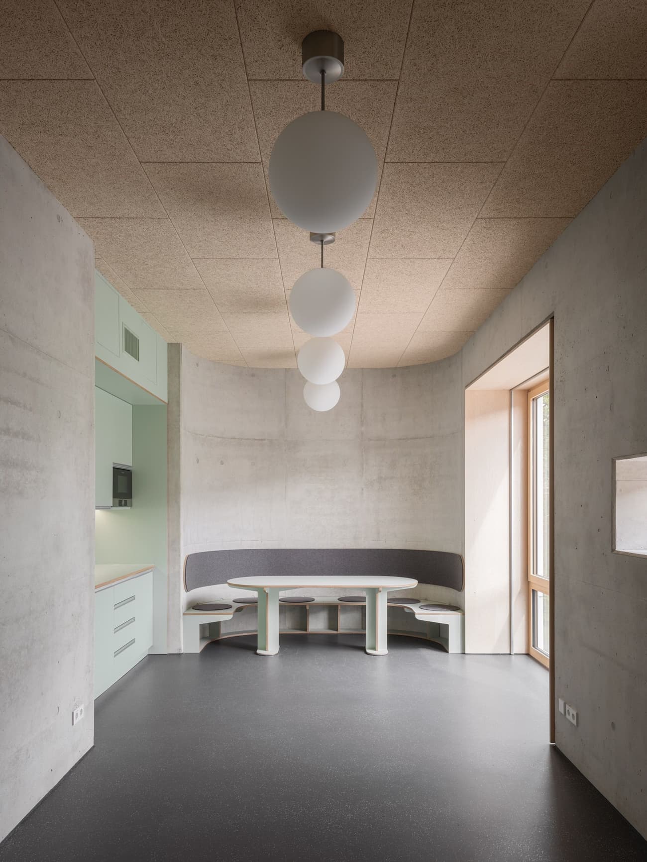Architectural project - exposed concrete, modern dining, round table, pendant lights, soft furnishings, mint green, corner seating, cork ceiling, globe lights, sleek cabinetry, integrated kitchen, chic nook, cork, cozy interior, natural materials, soft lighting, natural light, sustainable design, functional space, daylight, concrete, eco friendly, simple design, contemporary design, Scandinavian style, neutral palette, neutral colors, pastel green, window view, wood detailing, plaster, cozy nook, Nordic vibe, peaceful vibe, residential space, bright interior, minimalist kitchen, modern kitchen, breakfast nook, compact kitchen, calm atmosphere, compact living, home design, small kitchen, interior design, concrete wall, modern interior, muted tones, built-in bench, urban living, relaxation corner, architectural space