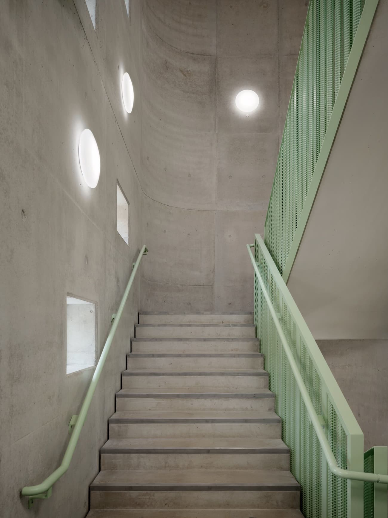 Architectural project - interior stair, industrial staircase, muted colors, mint green, concrete aesthetic, concrete stair, brutalist architecture, circular windows, green handrail, brutalism, indoor steps, building interior, modern stairwell, minimalist stairwell, wall lights, stair railing, mint accent, subtle color, structural clarity, simple stairs, round windows, soft lighting, raw materials, natural light, grey, green, functional space, daylight, industrial vibe, concrete, minimalist vibe, architectural lighting, minimalist architecture, contemporary design, neutral palette, minimalist design, geometric design, modern minimalism, white, sleek design, innovative materials, simple lines, clean design, concrete wall, efficient design, architectural feature