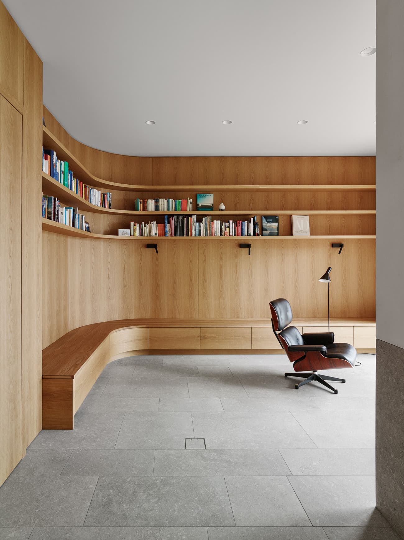 Architectural project - natural materials, soft lighting, timber, grey, natural light, inviting atmosphere, warm tones, neutral tones, functional space, daylight, light wood, natural wood, modern design, simple design, contemporary design, Scandinavian style, clean lines, elegant design, stone, wood shelves, relaxing space, cozy corner, reading nook, built-in shelves, home office, minimalist space, timber shelving, book nook, study area, reading area, comfortable chair, lounge chair, Eames chair, curved wood, timber panel, integrated lighting, grey floor, stone floor, minimalist aesthetic, home library, residential space, multifunctional room, ergonomic seating, minimal clutter, work from home, study zone, bright interior, modern furniture, light timber