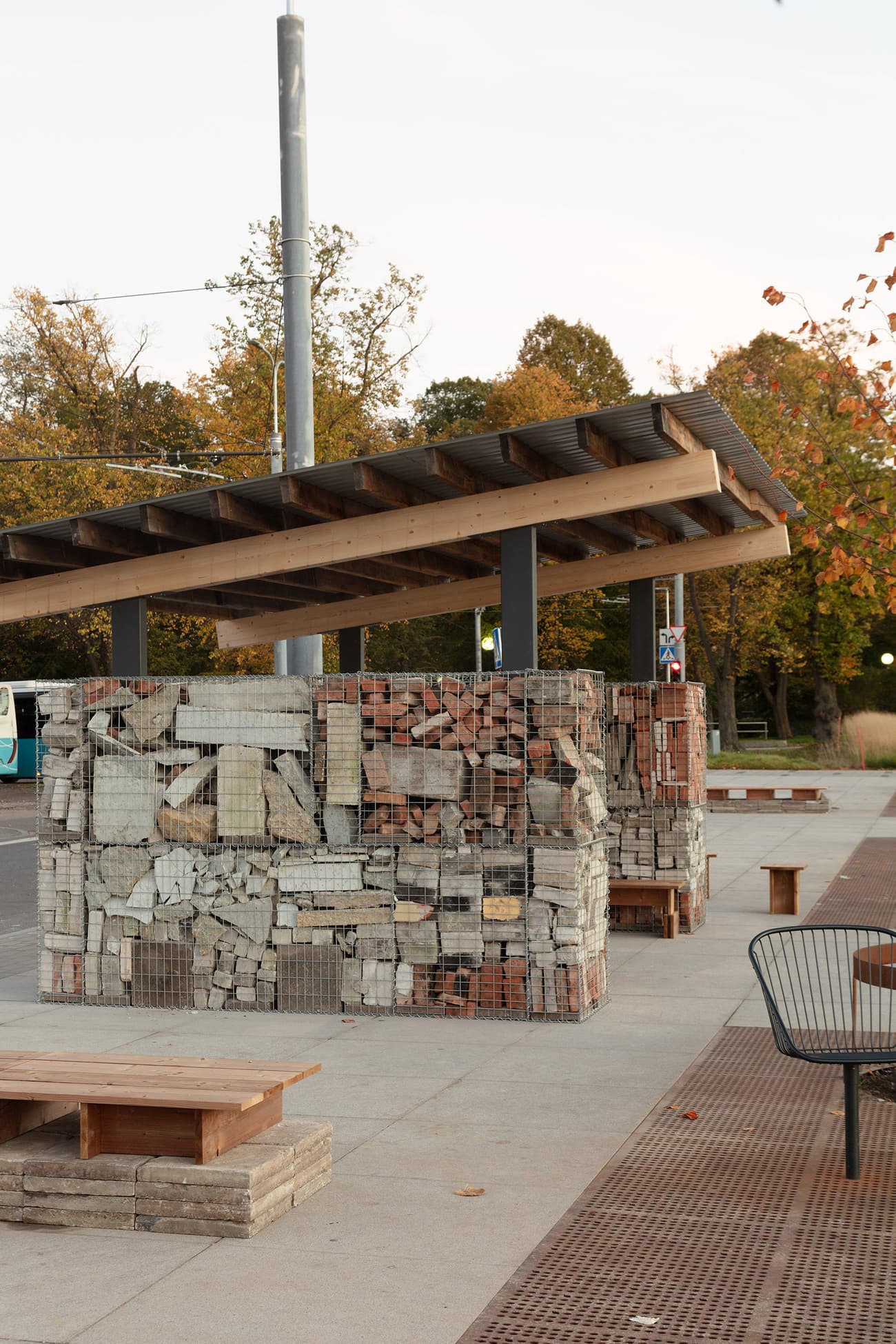Architectural project - pedestrian area, urban renewal, rustic bench, urban landscaping, open air space, public seating, gabion wall, concrete bench, public bench, street design, exposed bricks, fall setting, autumn scene, seating arrangement, concrete surface, recycled aggregates, cityscape, outdoor space, sustainable design, reclaimed materials, recycled materials, warm tones, eco design, community space, grey, urban environment, industrial vibe, gathering space, brown, eco friendly, natural setting, rustic vibe, contemporary design, minimalist design, outdoor seating, steel structure, urban design, street furniture, timber roof, wood roof, simple construction, outdoor gathering, slatted roof, green design, modern urban, outdoor bench, brick texture, red
