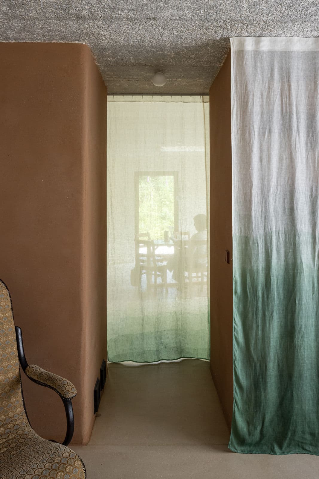 Architectural project - passageway, curtain divider, rustic interior, woven chair, concealed room, hidden room, fabric curtain, ombre curtain, green curtain, beige floor, vintage vibe, homely atmosphere, transitional space, relaxed corner, warm interior, intimate space, natural materials, soft lighting, serene environment, fabric, natural light, warm tones, green, neutral tones, daylight, beige, brown, concrete, eco friendly, organic design, calming space, earthy texture, sustainable home, textured wall, plaster wall, plaster, small space, cozy nook, private nook, rustic warmth, compact design, inviting space, earthy tones, relaxing space, cozy corner, lounge chair, cottage style, concrete floor, home interior, residential interior