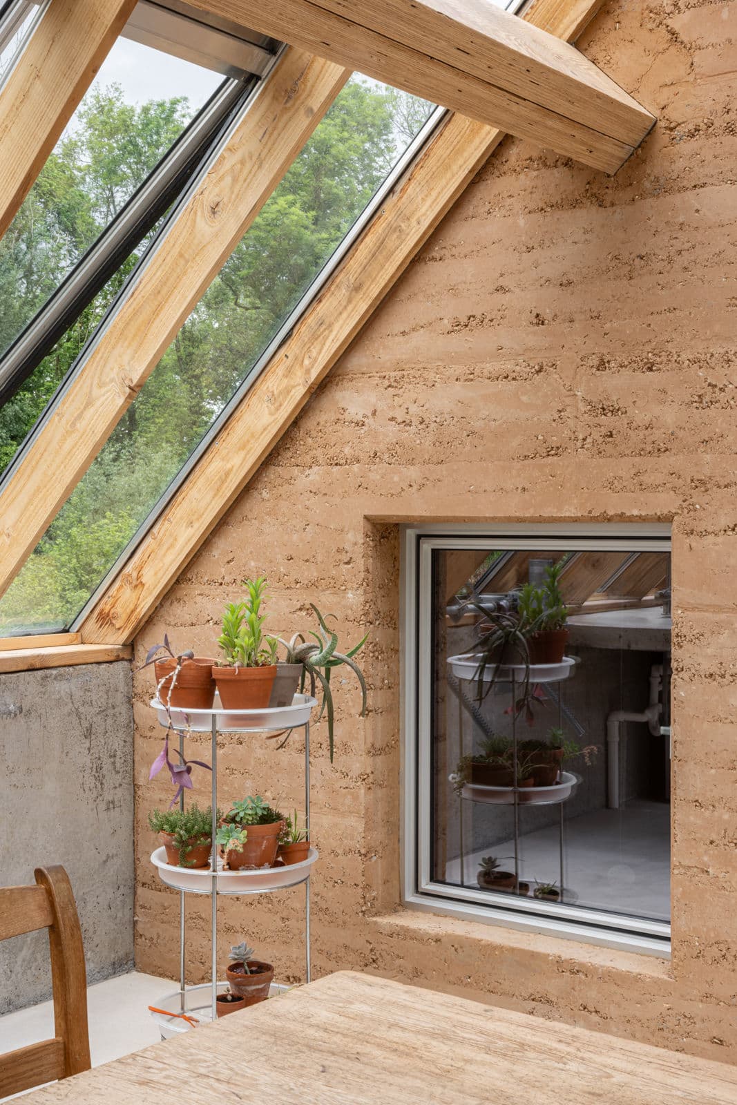 Architectural project - earth wall, attic space, loft conversion, potted plants, ceiling window, slanted roof, green view, terracotta pots, plant shelf, cozy roofspace, warm interior, natural materials, wood, timber, timber frame, natural light, sustainable design, warm tones, neutral tones, exposed beams, daylight, light wood, rustic vibe, wood beams, timber beams, eco materials, rammed earth, rammed earth wall, earth tones, biophilic design, eco home, indoor outdoor, window view, textured wall, plaster wall, energy efficient, cozy nook, rustic style, wooden table, inviting space, minimalist decor, earthy tones, natural texture, terracotta, green design, passive solar, sustainable living, simple furniture, connection to nature, sloped ceiling, skylight, indoor plants, vertical garden
