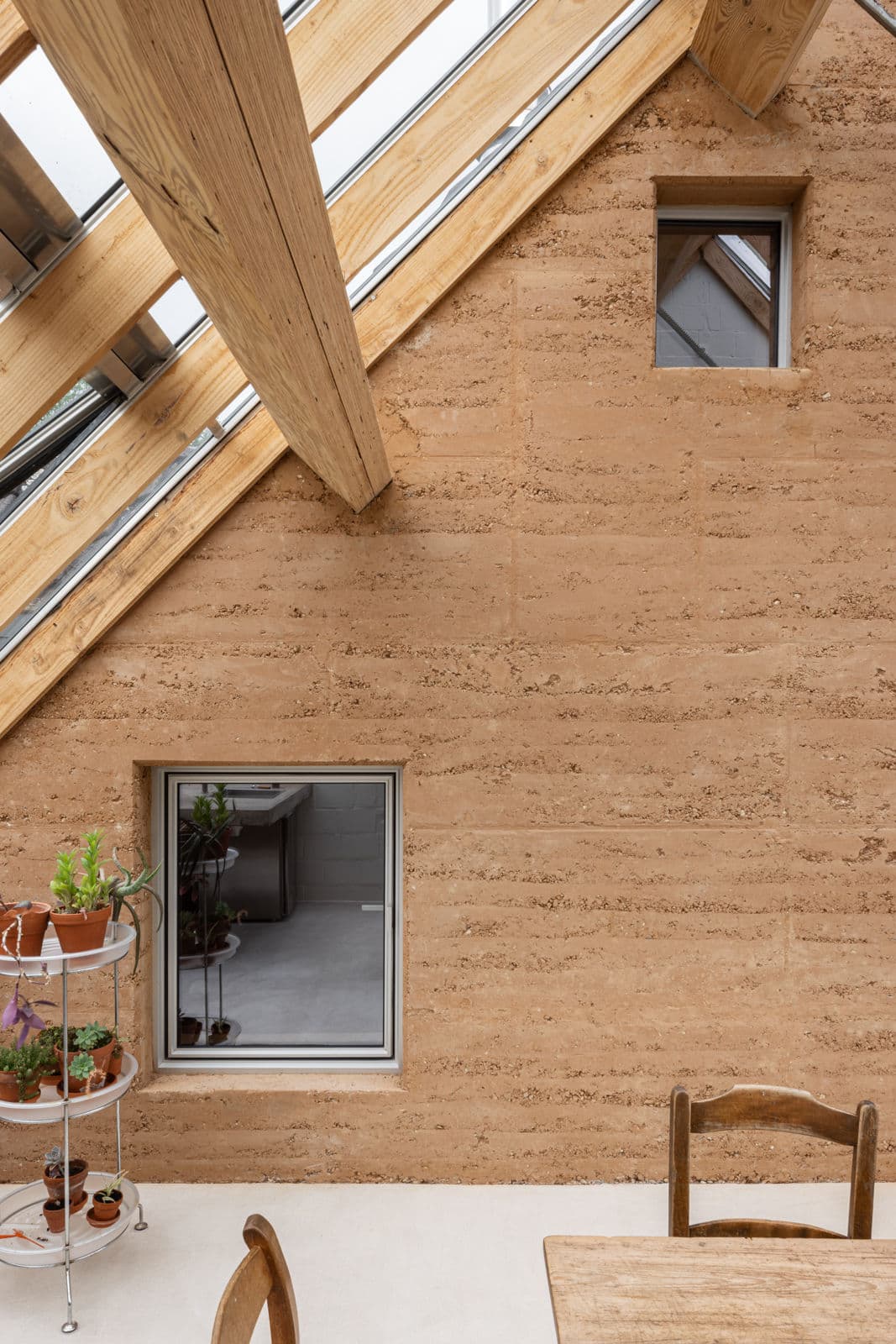 Architectural project - bio-based materials, wooden chairs, home dining, terracotta pots, plant shelf, farmhouse modern, clay tones, Scandinavian cabin, warm interior, natural materials, architectural detail, wood, timber, natural light, sustainable design, warm tones, eco design, green building, neutral tones, exposed beams, daylight, beige, light wood, compact space, sunlight, timber construction, rustic vibe, wood beams, timber beams, rustic modern, neutral palette, multifunctional, ecological design, rammed earth, rammed earth wall, low carbon, earth tones, passive house, eco friendly home, indoor outdoor connection, Nordic style, wood frame, rustic style, dining area, sloped roof, pitched roof, natural aesthetics, terracotta, cozy atmosphere, relaxing space, bright space, earthy textures, skylight, indoor plants, natural finishes
