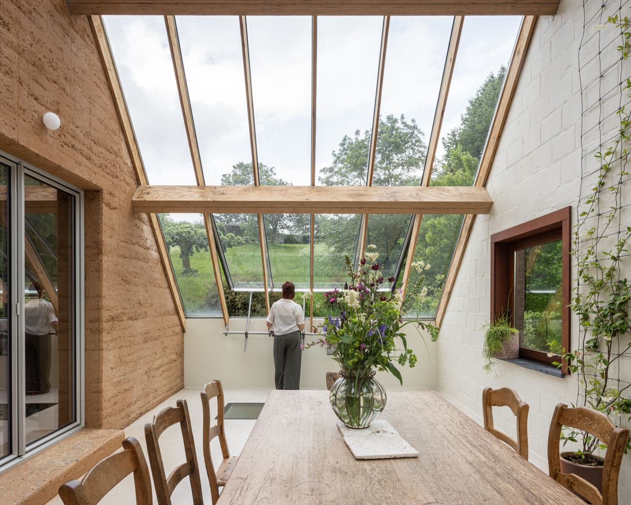Architectural project - home dining, indoor garden, glass roof, plant decor, rammed earth walls, white brick, rustic dining, farmhouse interior, rustic chairs, natural materials, serene environment, wood, timber, natural light, passive design, eco-conscious, sustainable design, glass, warm tones, daylight optimization, eco friendly, wooden beams, natural textures, rustic charm, Scandinavian style, minimalist style, cozy space, earth tones, modern rustic, biophilic design, nature view, white walls, green landscape, light filled, white, inviting home, warm ambiance, open layout, open plan, dining area, wooden table, earthy tones, brick, garden view, simple decor, nordic influence, airy atmosphere, indoor outdoor flow, rural retreat, skylight, vaulted ceiling