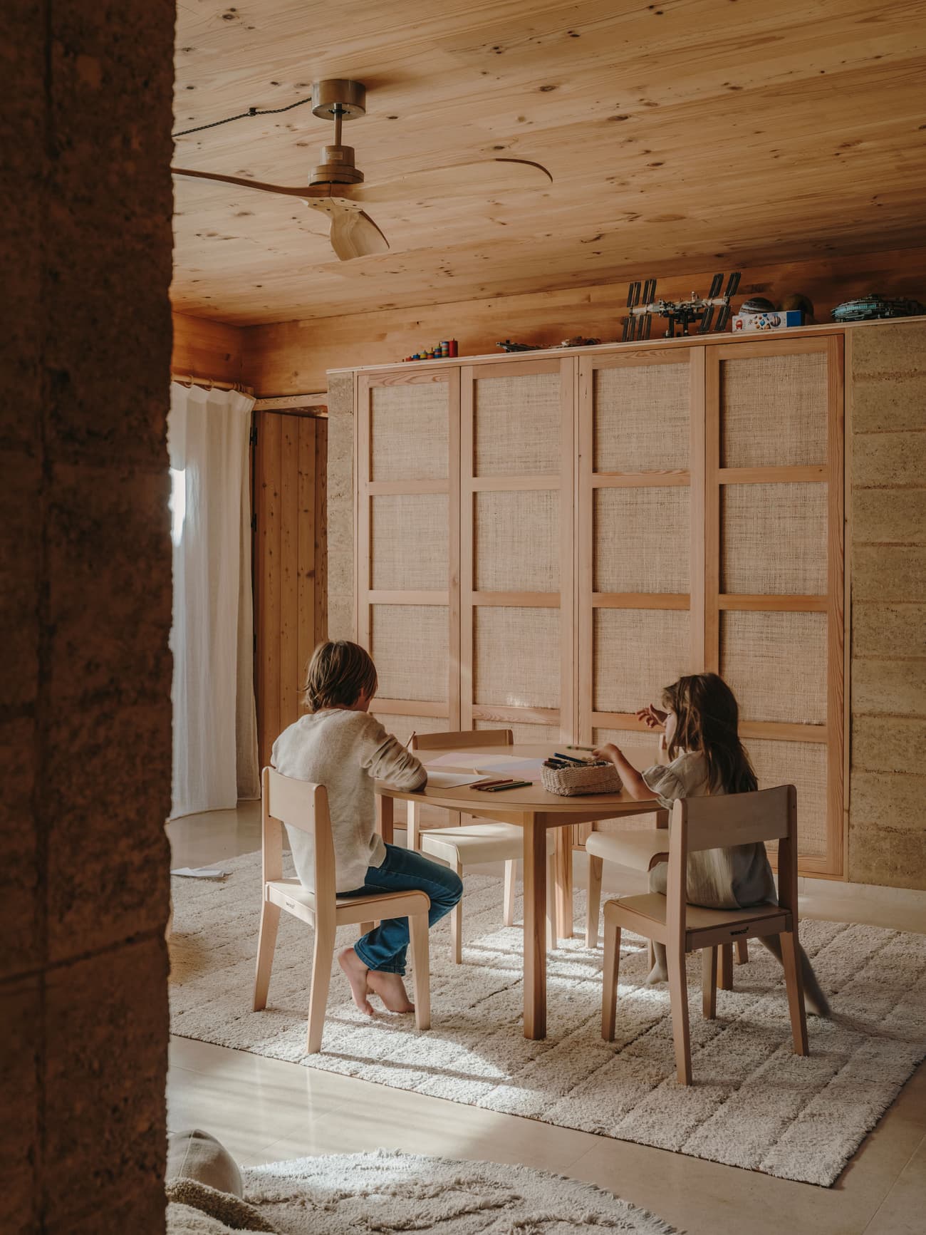 Architectural project - cozy living, home decor, minimalist furniture, calm environment, ceiling fan, small space living, woven details, kids area, floor rug, plush rug, textured fabric, natural materials, wood, timber, raw materials, natural light, warm tones, eco design, neutral tones, daylight, wooden ceiling, beige, brown, light wood, multifunctional space, honey wood, rustic modern, intimate setting, Scandinavian style, relaxed atmosphere, timber panels, modern rustic, sustainable home, Nordic design, cozy vibe, inviting home, casual living, timber interior, family space, living area, wooden table, textured walls, warm vibe, seating area, wood ceiling, interior design, simple furniture, home study, wood interior