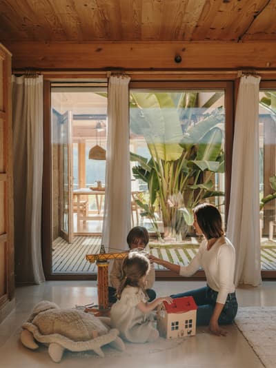 Architectural project - floor to ceiling windows, home decor, tropical plants, children playing, cozy environment, wooden toys, decoration, curtain, warm interior, wood, timber, relaxed vibe, natural light, sustainable design, glass, warm tones, neutral tones, exposed beams, daylight, beige, light wood, honey wood, Scandinavian style, biophilic design, large windows, indoor outdoor, white, Nordic design, eco friendly home, inviting home, timber ceiling, garden view, minimalist aesthetic, calm atmosphere, earthy textures, comfortable living, wood ceiling, family room, playful space, wooden furniture, sliding glass doors, play area, living space