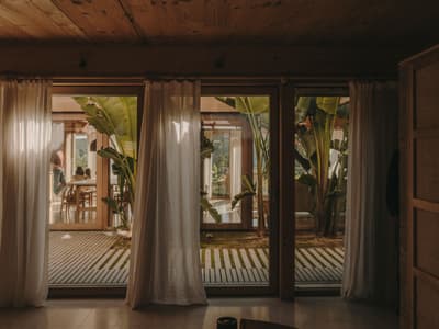 Architectural project - natural retreat, indoor courtyard, tropical house, plant-filled, open doors, sliding glass, houseplants, tropical vibe, social area, warm interior, natural materials, wood, timber, natural light, bohemian style, outdoor living, sustainable design, glass, warm tones, green, neutral tones, wooden ceiling, beige, brown, wood beams, natural textures, private retreat, intimate setting, biophilic design, eco home, large windows, indoor outdoor, cozy vibe, inviting home, timber house, rustic style, relaxed living, casual living, family space, earthy tones, nature-inspired, greenery, lush garden, garden view, wooden house, eco retreat, seamless transition