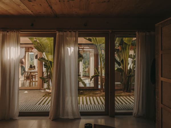 Architectural project - natural retreat, indoor courtyard, tropical house, plant-filled, open doors, sliding glass, houseplants, tropical vibe, social area, warm interior, natural materials, wood, timber, natural light, bohemian style, outdoor living, sustainable design, glass, warm tones, green, neutral tones, wooden ceiling, beige, brown, wood beams, natural textures, private retreat, intimate setting, biophilic design, eco home, large windows, indoor outdoor, cozy vibe, inviting home, timber house, rustic style, relaxed living, casual living, family space, earthy tones, nature-inspired, greenery, lush garden, garden view, wooden house, eco retreat, seamless transition
